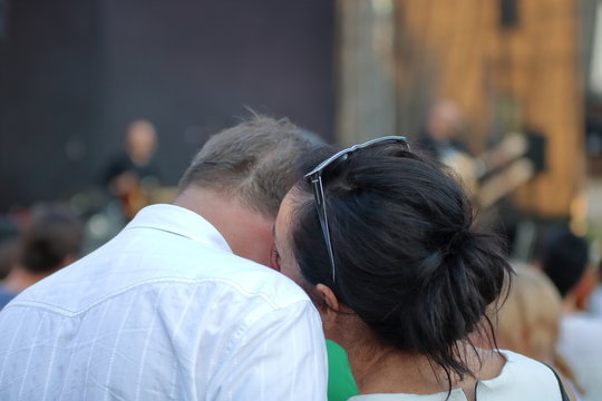 Dwoje zakochanych ludzi, mężczyzna i kobieta, w dojrzałym wieku, stoją tyłem (g&oacute;rne części ciała), eleganccy, pani przytula się i delikatnie całuje ukochanego, on nachyla do niej głowę, w tle ulica