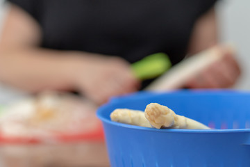 Two peeled pieces of white asparagus in front of a woman who is peeling white asparagus in the kitchen