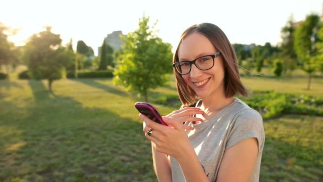 Smiling attractive girl chatting on her smartphone with friend browsing reading news. Sunlight sunbeams.