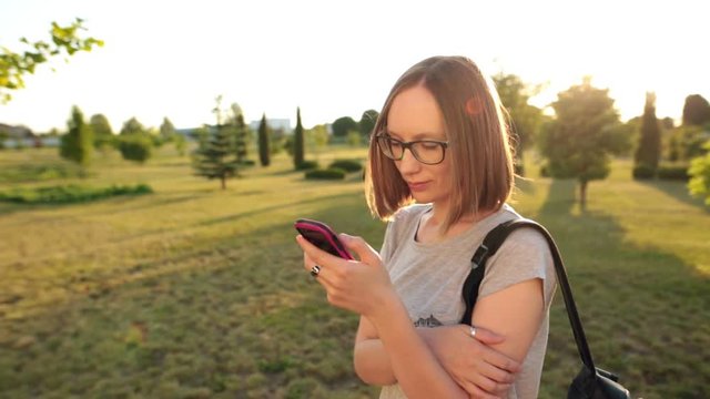 Casual girl in glasses chatting uses smartphone. Evening sun is shining.