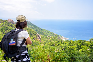 Women tourist with a backpack wear cap standing look at beautiful nature landscape blue sea under the summer sky from high scenic viewpoint at Koh Tao in Surat Thani, Thailand