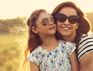 Happy fashion kid girl embracing her mother in trendy sunglasses and looking on nature background....