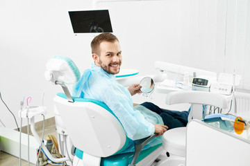 Young male client is smiling to the camera while checking his teeth in the mirror in a modern white dentist cabinet.