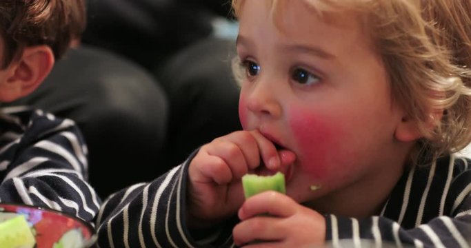 Cute adorable child starring at screen while chewing and eating cucumber. Candid real life authentic baby with red cheaks