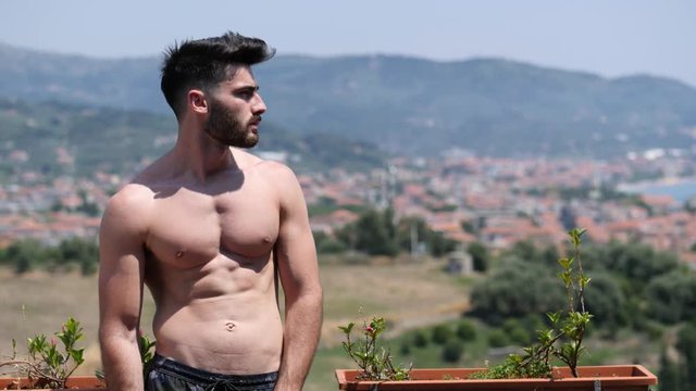 Handsome shirtless muscular young man outdoor on a balcony or terrace, wearing only shorts, looking away