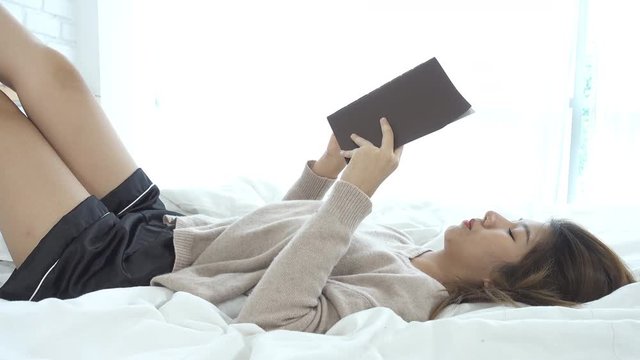 Lifestyle happy young Asian woman enjoying lying on the bed reading book pleasure in casual clothing at home. Relaxing lifestyle woman concept.