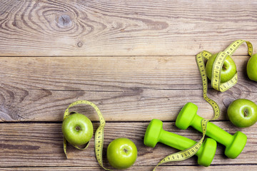 Green dumbbells and green apples with measuring tape and copy space on old wooden background. Sport, fitness and diet concept.
