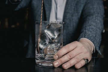 An underexposed image of barman's hands stirring ice in a mixing glass. Film grain effect.