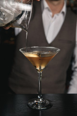 A vertical image of barman's hands, pouring a cocktail from a mixing glass into a martini glass