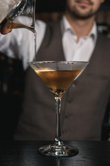 A vertical image of barman's hands, pouring a cocktail from a mixing glass into a martini glass