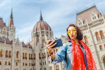 Fototapeta na wymiar Happy young asian woman tourist making selfie photo by using her smartphone standing in front of the old bridge in Budapest city with view of the Parliament Building