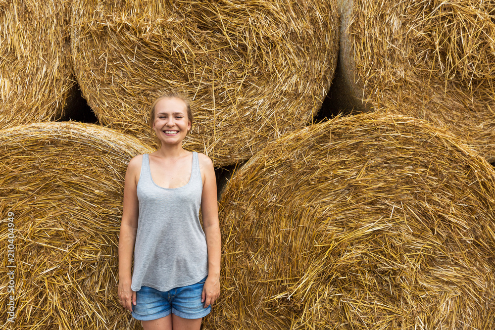 Wall mural a smiling young woman with blond hair stands on the background of haymaking.