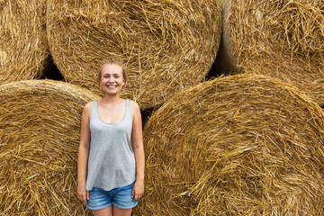 A smiling young woman with blond hair stands on the background of haymaking.