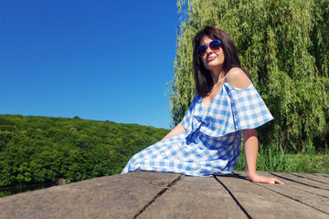 Young woman in sunglasses sits on the bridge near the river on a sunny day.