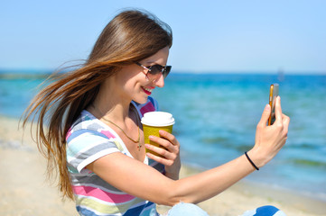 Young beautiful girl with glasses and a cup of coffee makes selfie on the seashore