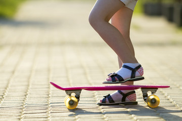Child slim legs in white socks and black sandals on plastic pink skateboard on bright sunny summer...