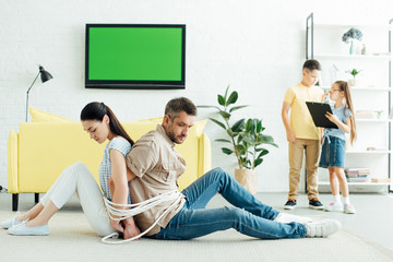 parents sitting tied with rope on floor and kids holding clipboard at home, parenthood concept