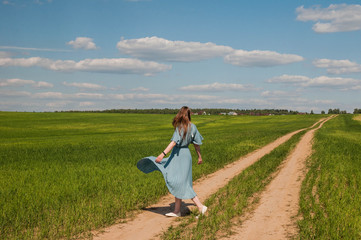 Naklejka na ściany i meble Beautiful young romantic girl outdoor in a blue dress with long hair enjoying nature, walking, spinning and dancing on the green field in the village