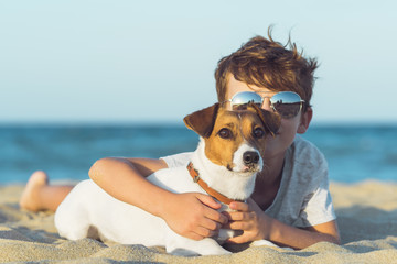 Happy boy hugging his dog breed Jack russell at the seashore against a blue sky close up at sunset. Best friends rest and have fun on vacation, play in the sand against the sea - obrazy, fototapety, plakaty