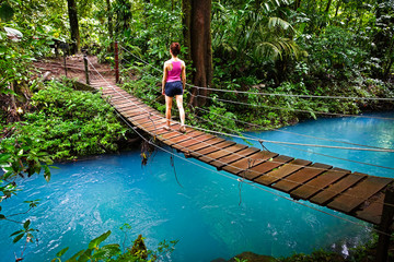 Young woman crossing a hanging bridge in the Jungle of Costa Rica. The river (rio celeste in...