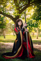 The Queen with a sword in her hands. Beautiful girl in the crown. Fantasy. Medieval black and red dress with gold embroidery.