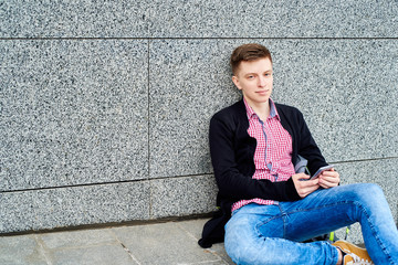 Cheerful young man in plaid shirt and jacket sitting under stone wall and using cellphone outdoors, free space. Student with mobile smart phone, free space