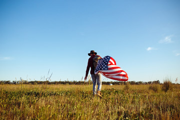 4th of July. American Flag.Patriotic holiday. Traveler with the flag of America. The man is wearing a hat, a backpack, a shirt and jeans. Beautiful sunset light. American style. 