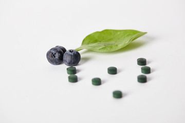 close up view of spirulina pills, blueberries and mint leaf on white background