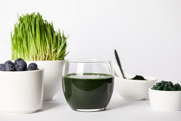 spirulina smoothie, spoon, cups with spirulina grass and blueberries, bowls with spirulina powder and spirulina pills on grey background