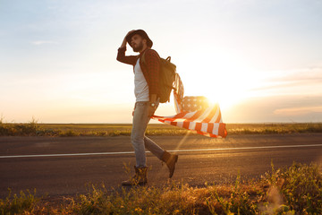4th of July. American Flag.Patriotic holiday. Traveler with the flag of America. The man is wearing a hat, a backpack, a shirt and jeans. Beautiful sunset light. American style. 