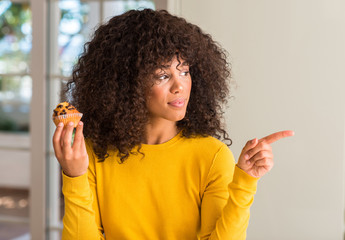 African american woman holding chocolate muffin very happy pointing with hand and finger to the side