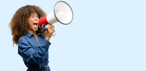 African american woman wearing blue jumpsuit communicates shouting loud holding a megaphone,...