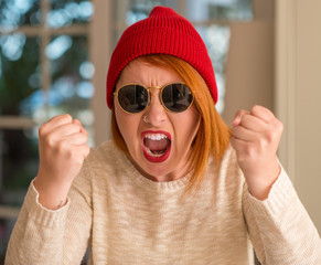 Stylish redhead woman wearing wool cap and sunglasses annoyed and frustrated shouting with anger, crazy and yelling with raised hand, anger concept