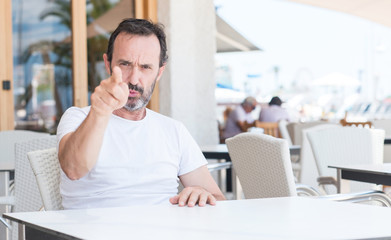 Handsome senior man sitting at terrace pointing with finger to the camera and to you, hand sign,...