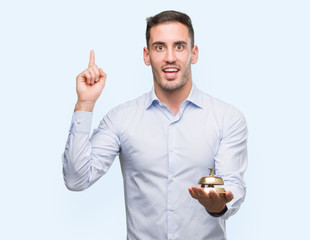 Handsome young man holding hotel ring bell surprised with an idea or question pointing finger with...