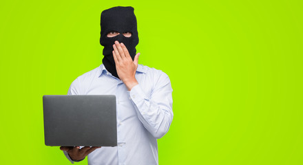 Business hacker man holding a computer laptop cover mouth with hand shocked with shame for mistake,...