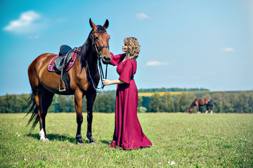 A beautiful girl in a long burgundy dress is standing in the field and holding a brown horse under the knots. Far away, another horse grazes.