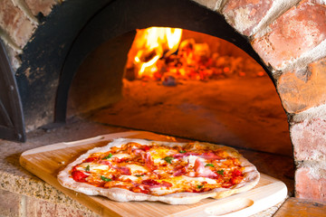 Rustic home-made pizza prosciutto baked in a wood fired brick oven with fire burning in the background