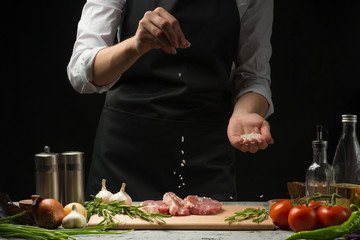 Chef salts steak grill pan. Preparing fresh beef or pork. Horizontal photo with a dark black background.