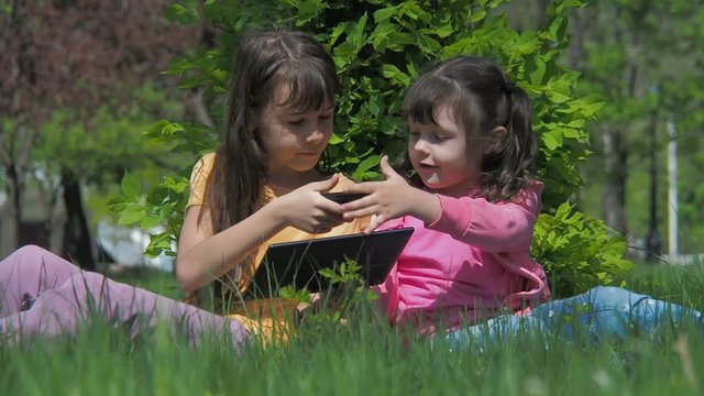 Children on the Internet. Sisters play on the tablet and phone sitting in the green grass.