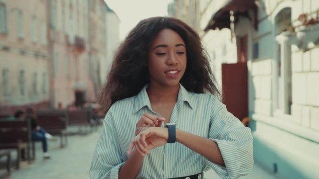 Smiling young woman walking to the work cross the city center, uses her smartwatch. Positive mood, contemporary technologies, gadgets. Successful lifestyle. Female portrait