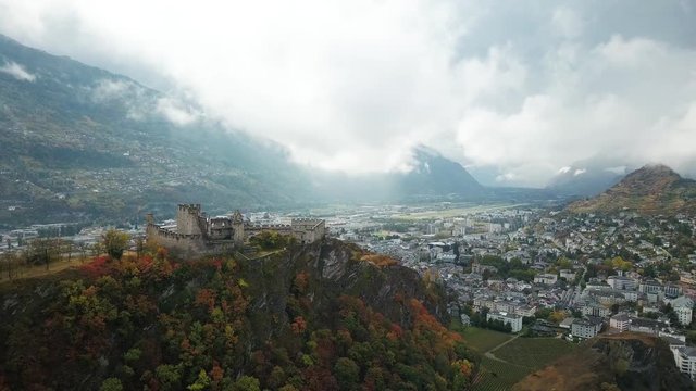 Sion Switzerland aerial view. Flying above hilltop castles in the Swiss alps in fall. Drone footage of cloudy mountains and village below in autumn.