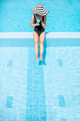 Young woman in black swimsuit and sun hat relaxing with cocktail in the swimming pool outdoors