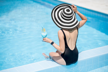Young woman in black swimsuit and sun hat sitting back with cocktail in the swimming pool outdoors