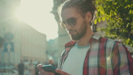 Young stylish young man with sunglasses standing using phone street texting casual clothing shirt city looking around sunny sunlight elegant trendy perfect look portrait slow motion close up