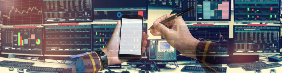 Double exposure of hand holding blank smartphone with Stock market quotes and chart in monitor computer room with business office equipments .business and money concept, panorama