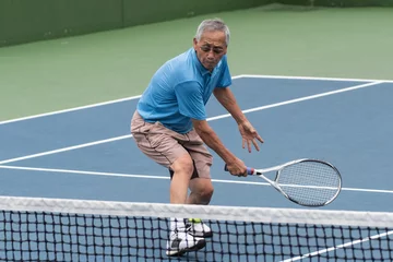 Tuinposter Healthy Chinese elderly man showing flexibility while reach for the low tennis backhand volley. © motionshooter
