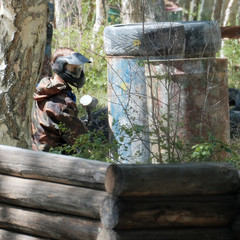 Obraz premium Extreme team sport. The player in the paintball looks out of hiding. Camouflage suit, protective mask and weapons. Tires, barrels, wooden fences in the forest.