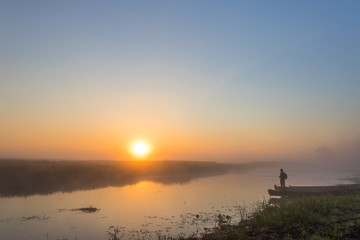 Foggy landscape with a fisherman standing on the river shore at dawn under the rising sun