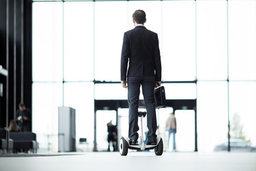 Back view of young businessman holding briefcase while moving forwards on hoverboard